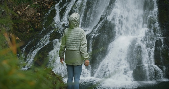 Woman traveler enjoying waterfall in the highlands of Austria. Traveling in the mountains, adventure and trip concept. Girl contemplates the beauty of nature in the forest.