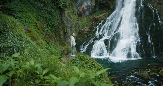 Woman enjoying the view of a powerful waterfall in green spring forest. Girl admires the beauty of nature. Hiking in the mountains. Hiking, adventure in trip concept.