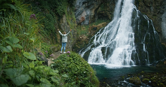 Girl admires the beauty of nature with her hands up. Woman traveler enjoys view of powerful waterfall in the highlands. Hiking in the mountains, adventure in trip concept.