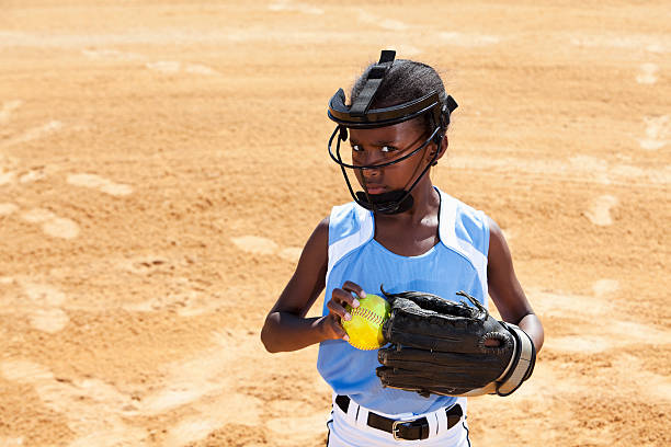 Softball player African American girl (9 years) playing softball, wearing face mask (standard safety gear required by many leagues). softball pitcher stock pictures, royalty-free photos & images