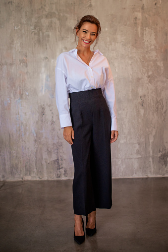 Full length of a beautiful brown-haired woman standing in front of a grey wall. Cheerful smiling female wearing white shirt and looking at camera. Copy space.