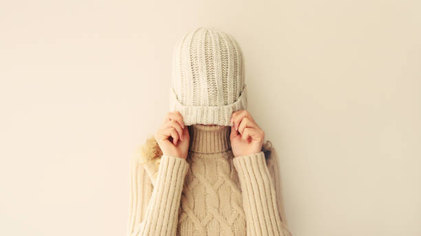 Cheerful funny woman having fun, pulling winter hat over her face and covering her eyes wearing warm soft knitted clothes, sweater on beige studio background