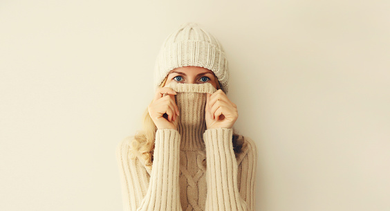 Winter portrait of woman freezing trying to warm up wearing warm soft knitted clothes, hat and sweater on beige studio background