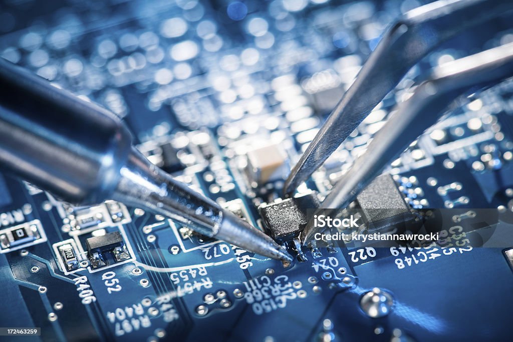 Soldering transistor on circuit board. Electronic technician soldering transistor using a soldering iron with the help of tweezers. Nice blue motherboard. Electronics Industry Stock Photo