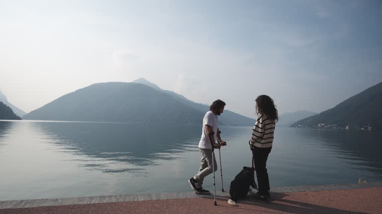 Man on crutches walks with his partner and their dog along boardwalk on lake