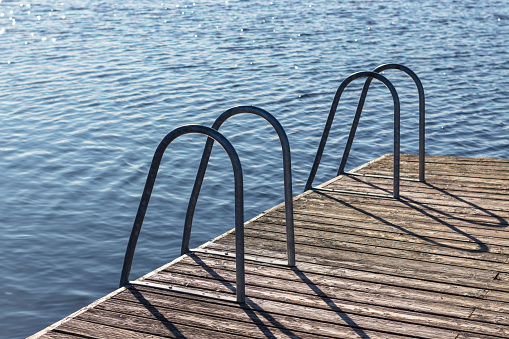 Metal dock ladder on wooden pear, blue water. Sea, ocean, river or lake. Summer swimming time.