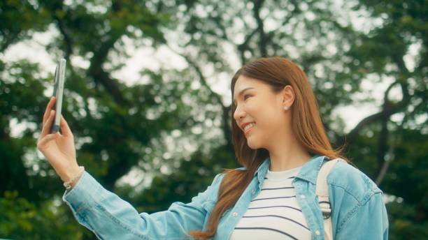 istock Cheerful Asian university student taking video call or selfie, walking under green trees on university park with happiness 1724639395