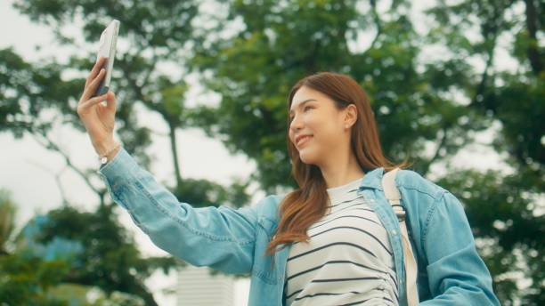 istock Cheerful Asian university student taking video call or selfie, walking under green trees on university park with happiness 1724639402