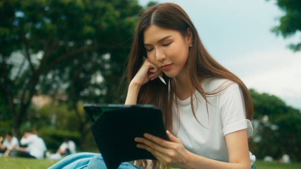istock Cheerful Asian female college student using digital tablet writing, reading, looking down in notes and sitting on grass in urban public park in city 1724639964
