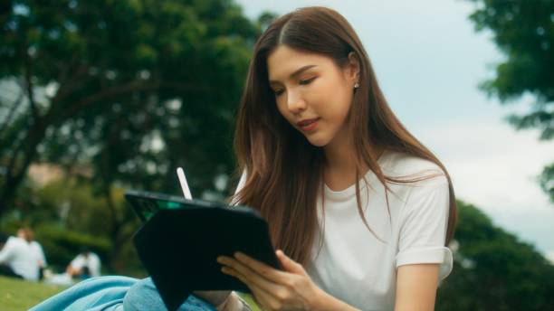 istock Cheerful Asian female college student using digital tablet writing, reading, looking down in notes and sitting on grass in urban public park in city 1724639985