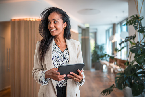 A mid-aged Hispanic businesswoman exudes confidence as she stands in a corporate office, armed with a digital tablet, effectively harnessing the potential of wireless technology in a modern business environment.