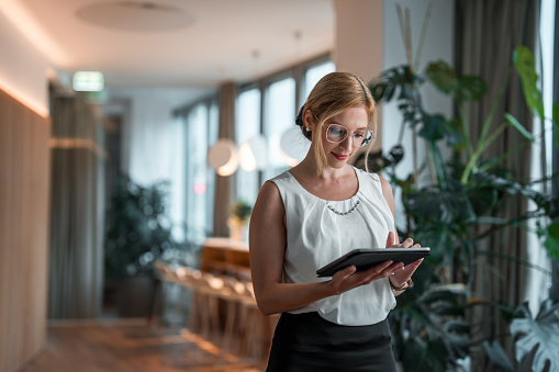 In a modern corporate environment, a knowledgeable mid-aged Caucasian female corporate employee stands out with her professionalism. She holds a digital tablet, employing digital tools adeptly to develop business strategies tailored to international clients.