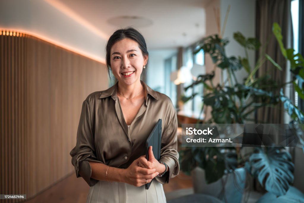 Smiling Female Korean Corporate Employee with Digital Device Positioned within a modern corporate environment, a mid-aged Asian businesswoman of striking beauty stands in her office, poised with a digital tablet, using it to execute digital tools and wireless technology to perfection in her strategic endeavors. Businesswoman Stock Photo