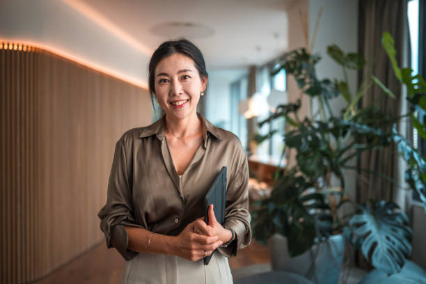 Smiling Female Korean Corporate Employee with Digital Device Positioned within a modern corporate environment, a mid-aged Asian businesswoman of striking beauty stands in her office, poised with a digital tablet, using it to execute digital tools and wireless technology to perfection in her strategic endeavors. businesswoman stock pictures, royalty-free photos & images