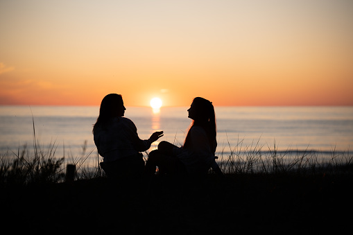 Silhouette of two female friends enjoying a conversation while sitting and watching the sunset on a sea beach. Back view