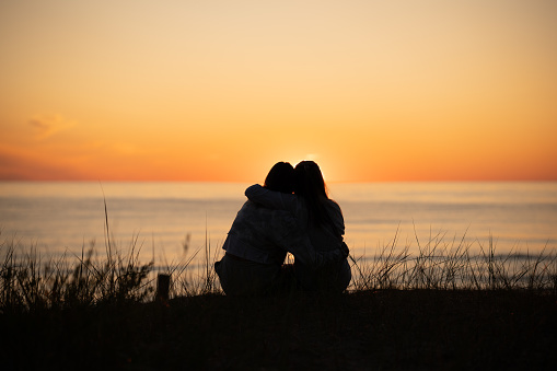 Silhouette of two female friends enjoying a conversation while sitting hugging and watching the sunset on a sea beach. Back view