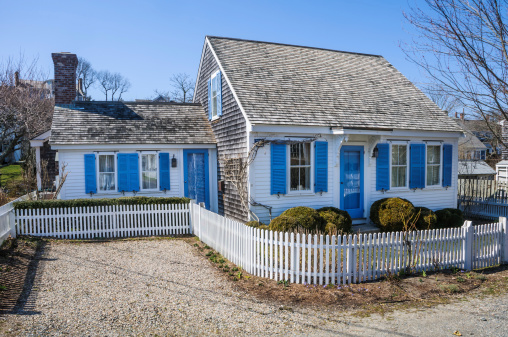 A low white picket fence surrounds the tiny house lot of this blue shuttered cottage at the tip of Cape Cod.