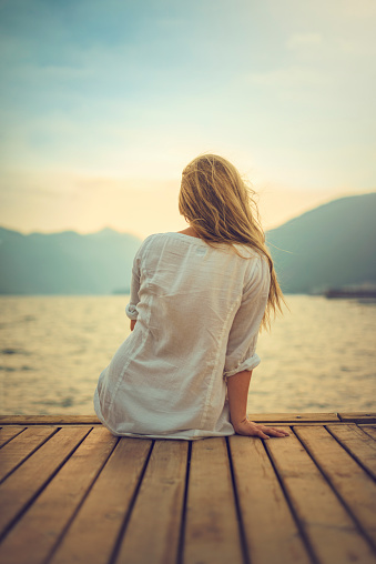 A young woman sits at the edge of a wooden dock.  She faces away from the viewer and looks at the view of a lake and distant mountains.  The woman wears a loose white top with the sleeves rolled up, and her long blonde hair is brushed forward over her right shoulder.  Before her is a lake with a lot of reflection from the sun.  In the distance, on the other side of the lake, are some out of focus mountains.  The sky above them is a bright white that fades to blue higher up.