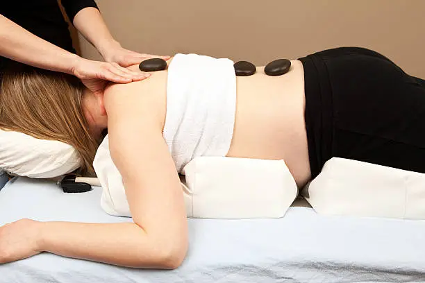 "A pregnant woman is receiving a prenatal massage from a female massage therapist. This type of massage is used to relieve tension, stress and discomfort during pregnancy. The pregnant woman is on a specialized bolster which provides space for her belly while on her stomach."