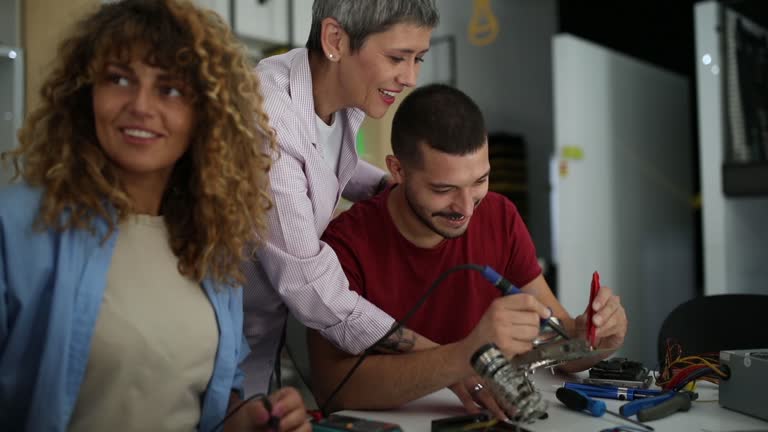 Carefree female teacher helps her students learn how to use a soldering iron, tweezers in an electronics class