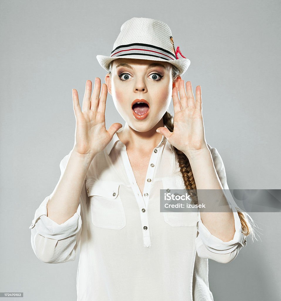 Young woman screaming, Studio Portrait Portrait of  young adult woman wearing hat, screaming at camera. Studio shot on grey background. Front View Stock Photo