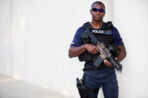 African American police officer (20s) wearing bullet proof vest, holding rifle.