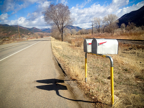 a pair of mailboxes on the side of a country road underneath a cloud filled blue sky with mountain ridges surrounding the valley landscape.  such beautiful nature scenery and communication americana can be found in the animas valley of durango colorado in the san juan range of the colorad rocky mountains.