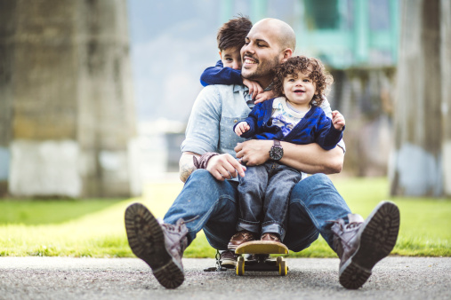 A Hispanic man with his two young sons riding on a skateboard.  The father is one son on his lap, and the other son is behind his father holding onto his neck.