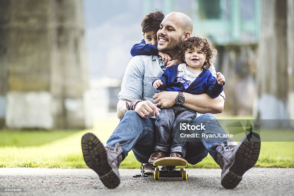 Dad on a skateboard at a park with two sons A Hispanic man with his two young sons riding on a skateboard.  The father is one son on his lap, and the other son is behind his father holding onto his neck. Father Stock Photo