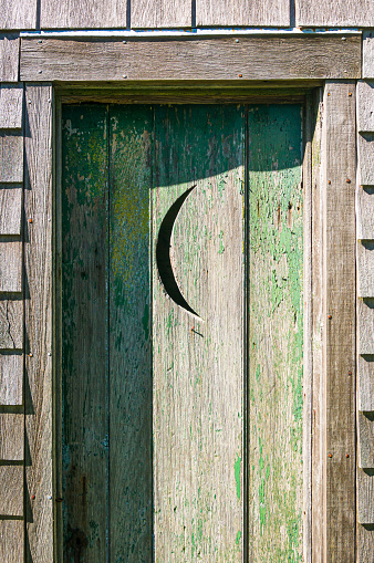 A rustic outhouse  with a green peeling paint on the door features a cresent moon on the door