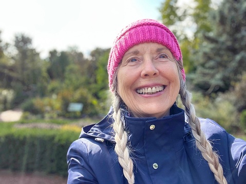 Close up portrait of cheerful senior woman in pink knit hat over braided hair on an outing on a beautiful autumn day.   Selfie