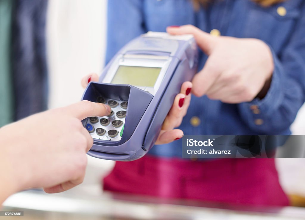 Finger entering pin code into credit card machine A woman types her pin code into a handheld credit card swiper.  Only the woman's right hand is visible as she enters the code.  The credit card swiper has a small screen and a shield over the 10-key numeric key pad to protect it from view.  The swiper is held by a woman standing behind a counter.  The woman wears a bright blue long-sleeved shirt and red pants, and she has bright red fingernails.  The woman holding the credit card swiper and the background behind her are all out of focus. ATM Stock Photo