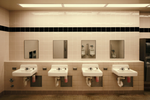 A sparse public men's restroom with sinks and mirrors against a wall. Urinals can be seen in the reflection of the mirrors.