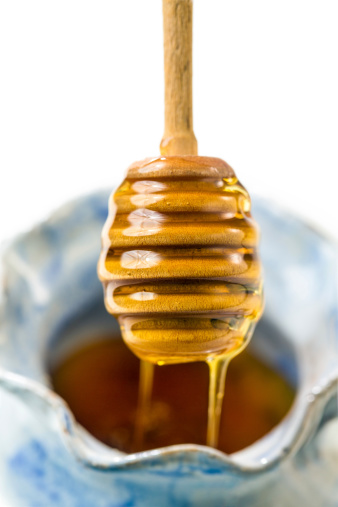 Sweet golden honey dripping from a honey dipper into ceramic honey pot on white background