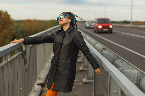 Woman with dreadlocks in black coat on bridge. Street style.