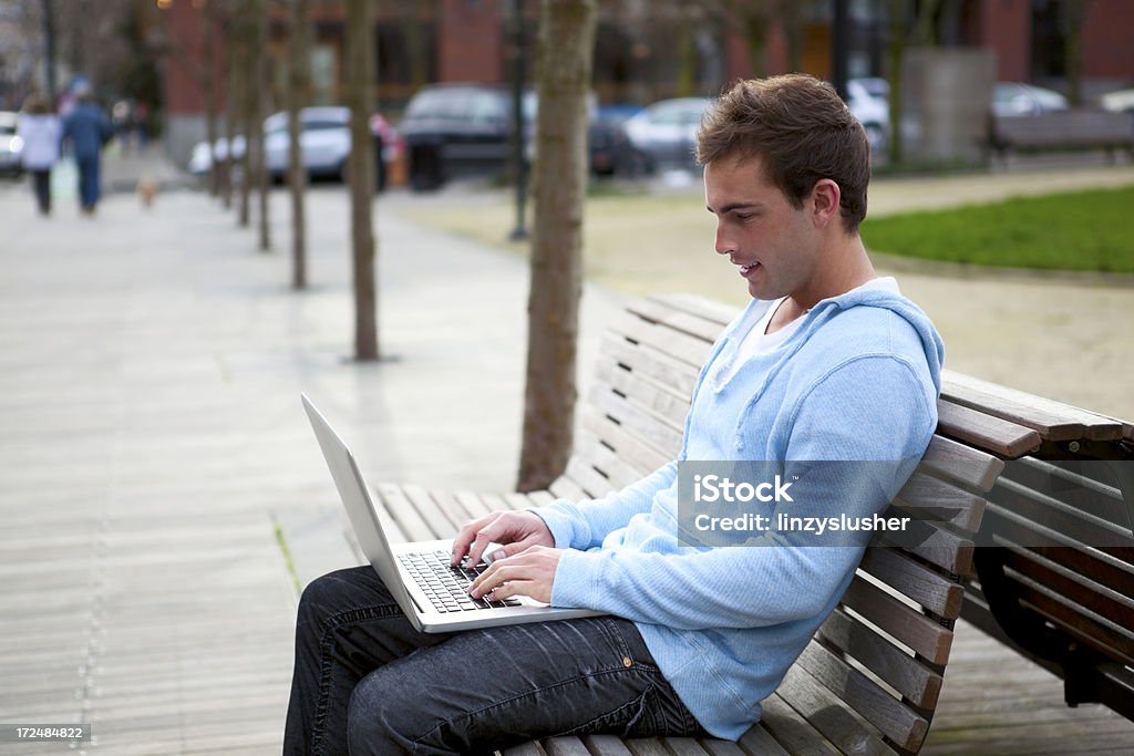 Atractiva joven hombre utiliza un ordenador portátil en un parque de banco - Foto de stock de Adulto joven libre de derechos