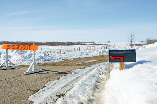 In early spring a  black rural mailbox along a rural countyr road with melting snow along the road's shoulders. Copy space on the mailbox. A sign shows the bridge is out on this road.