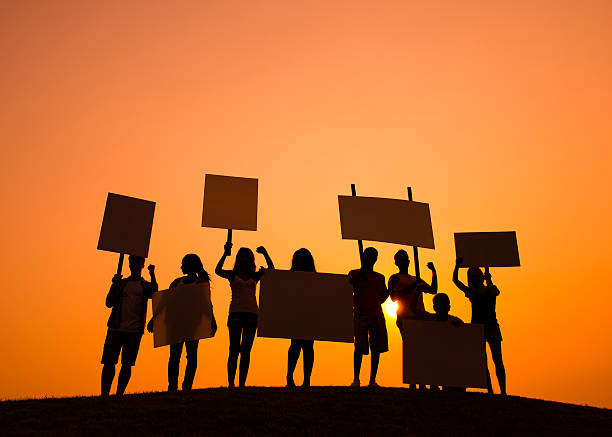 Silhouette of protestors holding signs against an orange sky A group of young people protesting at sunset/sunrise strike protest action photos stock pictures, royalty-free photos & images