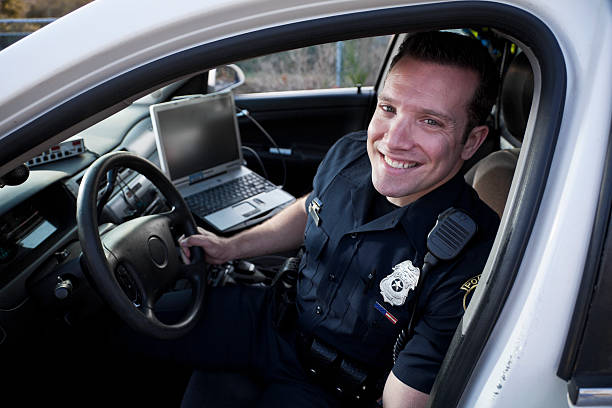Police officer sitting in cruiser Police officer (20s) sitting in police car, with laptop computer. police force smiling officer stock pictures, royalty-free photos & images