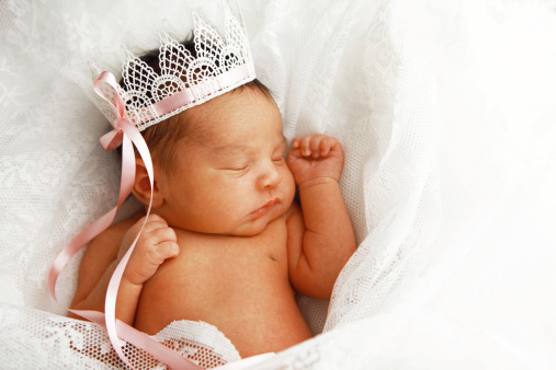 A portrait of a sleeping newborn baby girl wearing a pretty princess crown.  She is surrounded by pretty, white lace.