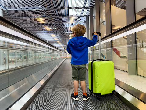 Little boy stand on moving walkway at the airport with green baggage suitcase view from behind