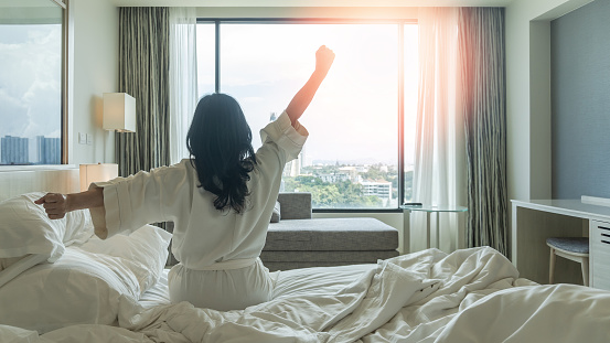 Hotel relaxation on lazy day with Asian woman waking up from good sleep on bed in weekend morning resting in comfort bedroom looking toward city view, having happy, work-life quality balance lifestyle