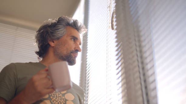 istock Mature man drinking coffee and looking through window blinds. 1724982241