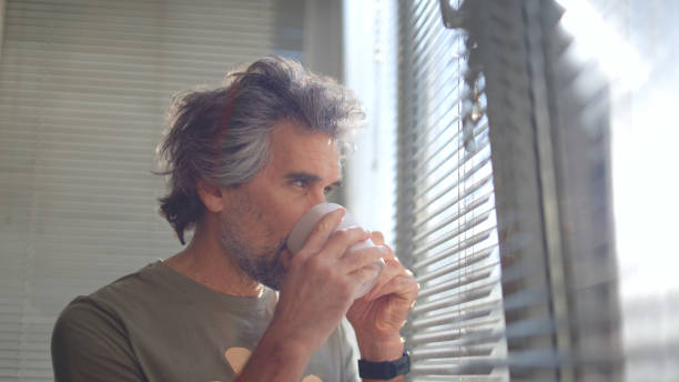 istock Mature man drinking coffee and looking through window blinds. 1724982251