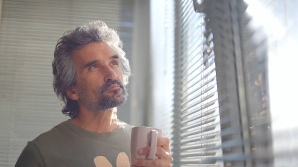istock Mature man drinking coffee and looking through window blinds. 1724982256