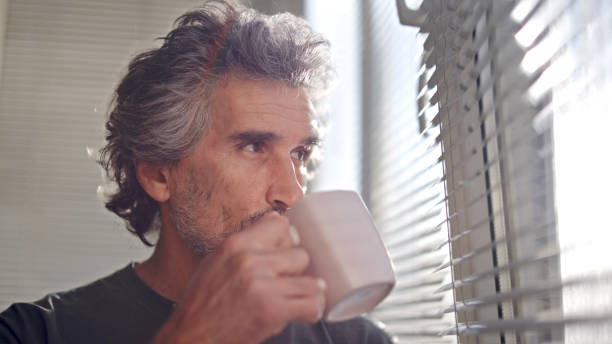 istock Mature man drinking coffee and looking through window blinds. 1724982263