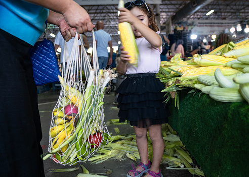Candid photo of mother and 5 years old daughter shopping in farmer's market. Mother is holding a reusable mesh bag full of vegetables and fruits. Shot under daylight in outdoor.