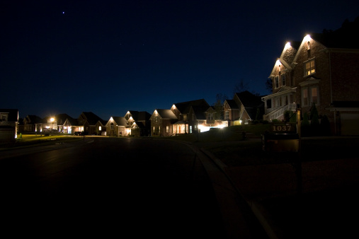 Neighborhood at night; houses all lit up with dramatic lighting. A starry and glowing sky silouhettes the rooftops.