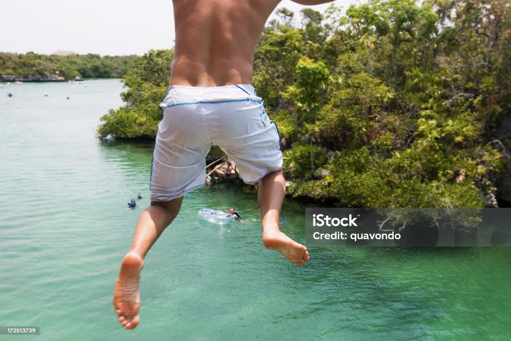 Back of Young Man Jumping off Cliff into River, Copyspace Boy jumping off a cliff into the river during summer. Copy space. Adolescence Stock Photo