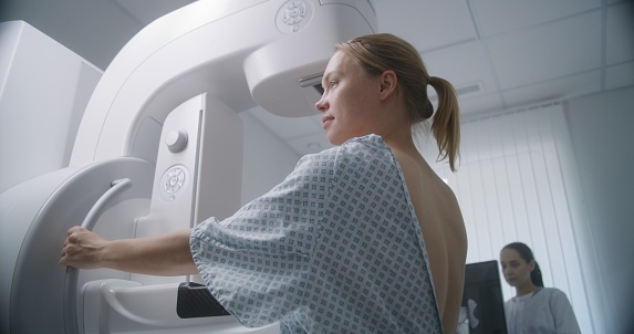 Caucasian adult woman stands in hospital radiology room. Female patient undergoing mammography screening procedure using digital mammogram machine. Breast cancer prevention. Modern clinic equipment.